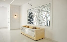 A beige sofa wrapped in plastic beneath a geometric mirror in a white room.