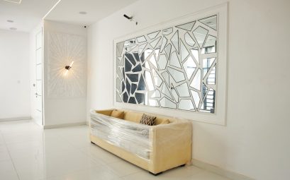 A beige sofa wrapped in plastic beneath a geometric mirror in a white room.