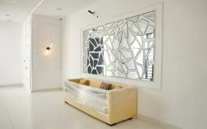 A beige sofa wrapped in plastic beneath a geometric mirror in a white room.