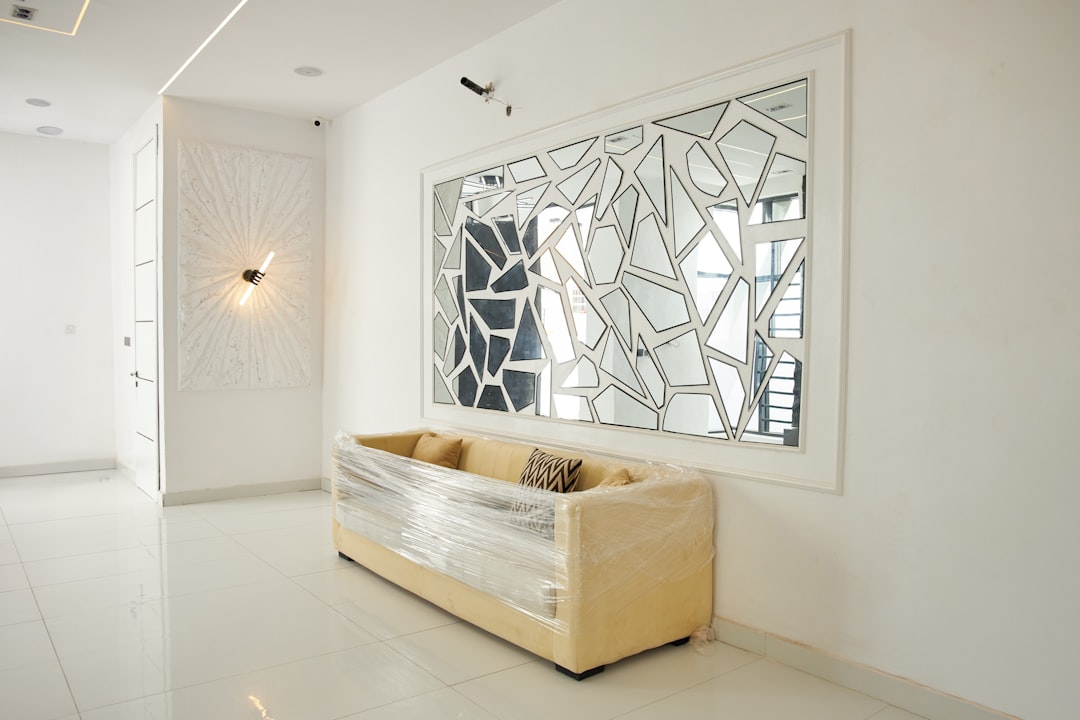 A beige sofa wrapped in plastic beneath a geometric mirror in a white room.